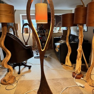May include: A tall, wooden floor lamp with a unique, organic shape. The lamp has a brown wooden base and a light brown shade. The lamp is lit and casting a warm glow.