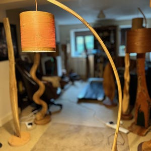 May include: A wooden floor lamp with a curved wooden arm and a cylindrical lampshade. The lampshade is made of a natural material and has a reddish-brown color.