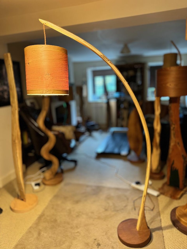 May include: A wooden floor lamp with a curved wooden arm and a cylindrical lampshade. The lampshade is made of a natural material and has a reddish-brown color.