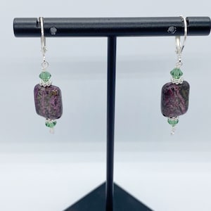 Puede incluir: Un par de pendientes con cierre de palanca. Cada pendiente presenta una piedra rectangular de color púrpura oscuro con detalles verdes y plateados. Cuentas de cristal verde se colocan encima y debajo de la piedra, conectadas por alambre de plata.