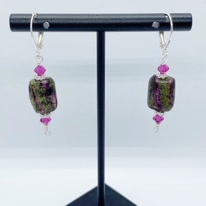 May include: Dangle earrings with olive green and purple rectangular stones. Each earring has a magenta crystal bead above and below the stone, with silver wire accents and lever-back closures.