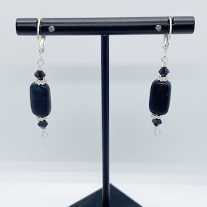 May include: A pair of black dangle earrings with a rectangular bead and small black crystal accents. The earrings are suspended from silver-colored lever-back closures and are displayed on a black jewelry stand.