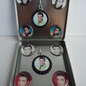 May include: A silver metal box with a hinged lid containing eight round button-shaped earrings with images of Elvis Presley. The earrings are silver with black accents and are displayed on a silver background.