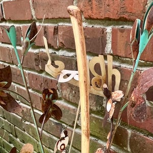 May include: A rustic outdoor scene featuring a light-colored ceramic pot holding metal floral sculptures and a wooden walking stick. The metal flowers have brown and teal accents. The background is a red brick wall.