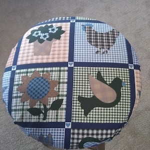 May include: A round blue, green, brown, and white fabric chair cushion with a patchwork design featuring a sunflower, a bird, a chicken, and a potted plant.