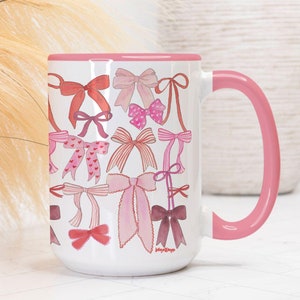 May include: A white ceramic mug with a pink rim and handle. The mug is decorated with a pattern of pink and red bows.