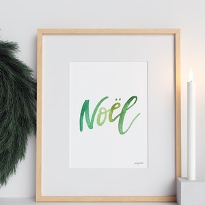 May include: A framed print with the word "No&euml;l" written in green watercolor lettering. The print is displayed on a white surface with two white candles on either side.