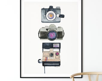 May include: A framed watercolor painting of three vintage cameras. The cameras are arranged vertically, with the oldest camera at the top and the newest camera at the bottom. The cameras are all different colors and styles, and they are all in focus. The background is white.