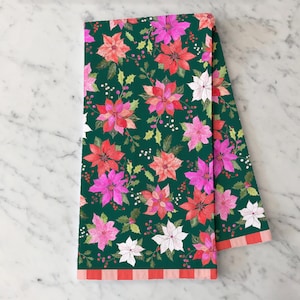 May include: Folded green kitchen towel with a floral pattern of red, pink, and white poinsettias and green leaves. The towel's border features a red and white striped design. The towel is folded and displayed on a marble surface.