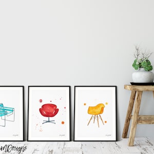 May include: Three framed watercolor illustrations of iconic chairs. The first chair is a blue and white lounge chair. The second chair is a red egg chair. The third chair is a yellow and brown Eames chair.