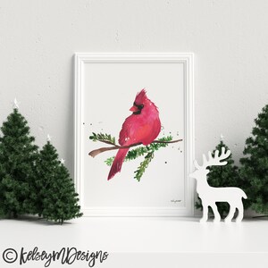 May include: A watercolor painting of a red cardinal perched on a branch with green leaves. The bird is facing left and has a black eye and beak. The painting is framed in a white frame.