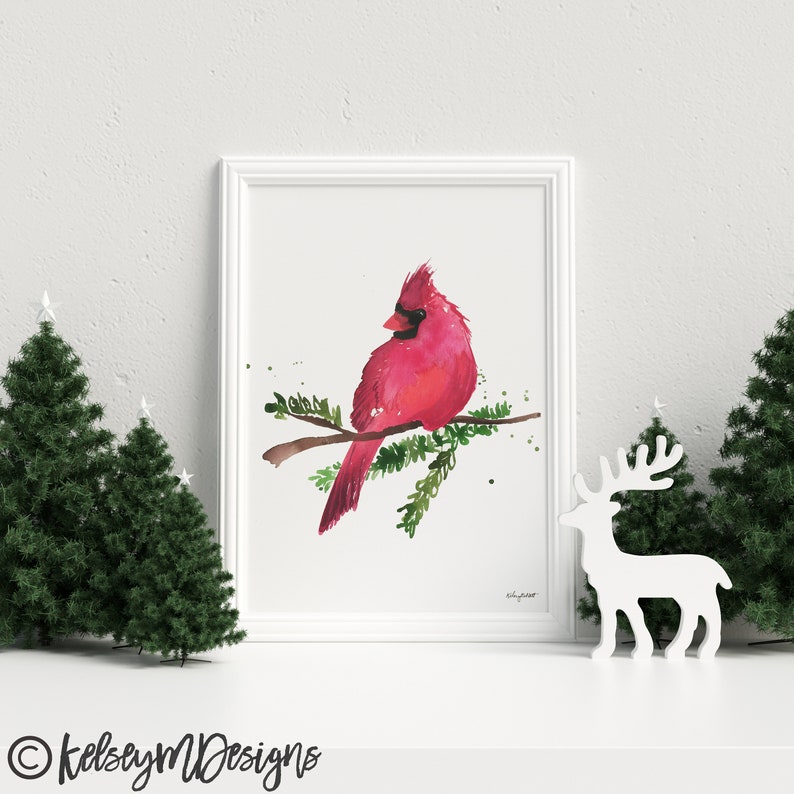 May include: A watercolor painting of a red cardinal perched on a branch with green leaves. The bird is facing left and has a black eye and beak. The painting is framed in a white frame.