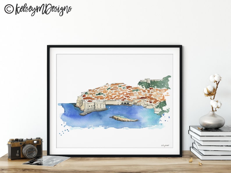 May include: Watercolor painting of a city on a peninsula with a blue ocean surrounding it. The city is made up of many buildings with red tile roofs. The painting is framed in a black frame.