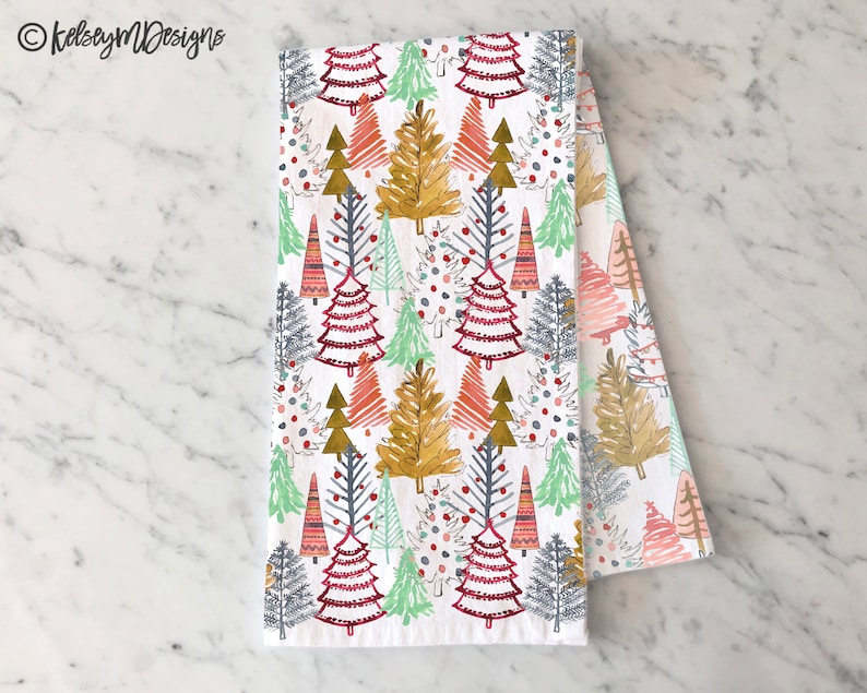 May include: White kitchen towel with a repeating pattern of colorful Christmas trees in shades of red, green, gold, and blue. The trees are stylized with various designs. The text "kelseymDesigns" is in the upper left corner.