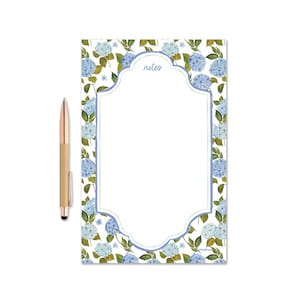 May include: A notepad with a floral hydrangea design and the word "notes" in script. The notepad has a light blue border and is accompanied by a gold pen with a black stylus tip. The notepad is approximately 8.5 inches tall.