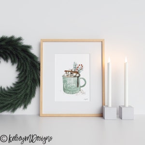 Puede incluir: Una impresi&oacute;n enmarcada con una ilustraci&oacute;n de acuarela de una taza verde llena de chocolate caliente, malvaviscos, crema batida y un palo de canela. El texto en la taza dice "Hot Cocoa with marshmallows served here".
