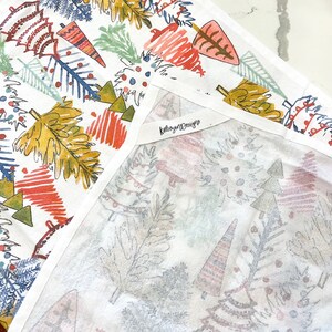 May include: White cotton tea towel with a colorful Christmas tree design. The trees are in shades of red, yellow, blue, and green. The towel has the text "kelseymdesigns" on a tag.
