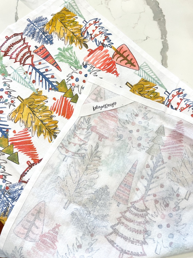 May include: White cotton tea towel with a colorful Christmas tree design. The trees are in shades of red, yellow, blue, and green. The towel has the text "kelseymdesigns" on a tag.