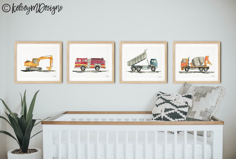 May include: Four framed watercolor prints of construction vehicles: an excavator, a fire truck, a dump truck, and a cement mixer. The prints are in light wood frames and are displayed above a white crib. The artist's name, kelseymdesigns, is in the top left corner.