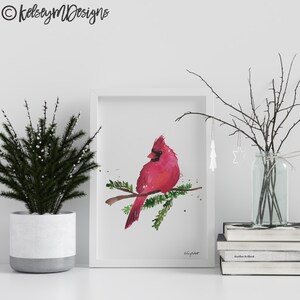 May include: A watercolor painting of a red cardinal perched on a branch with green leaves. The painting is framed in a white frame and is displayed on a white shelf with a small potted plant and a vase of branches.
