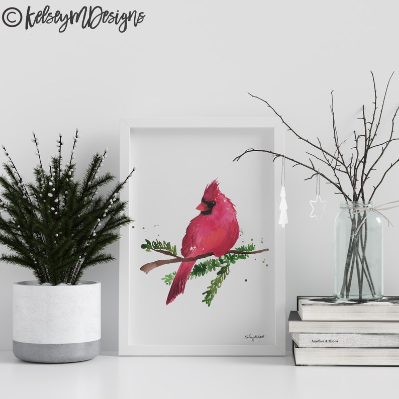 May include: A watercolor painting of a red cardinal perched on a branch with green leaves. The painting is framed in a white frame and is displayed on a white shelf with a small potted plant and a vase of branches.