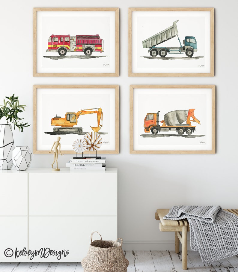 May include: Four framed watercolor prints of construction vehicles: a red fire truck, a blue dump truck, an orange excavator, and an orange cement mixer. Each print is in a light wood frame and hangs on a white wall.