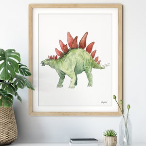 May include: Watercolor print of a green Stegosaurus with red plates, framed in a light wood frame. The artwork is on a white background. The artist's signature is visible. The print is displayed on a white surface.