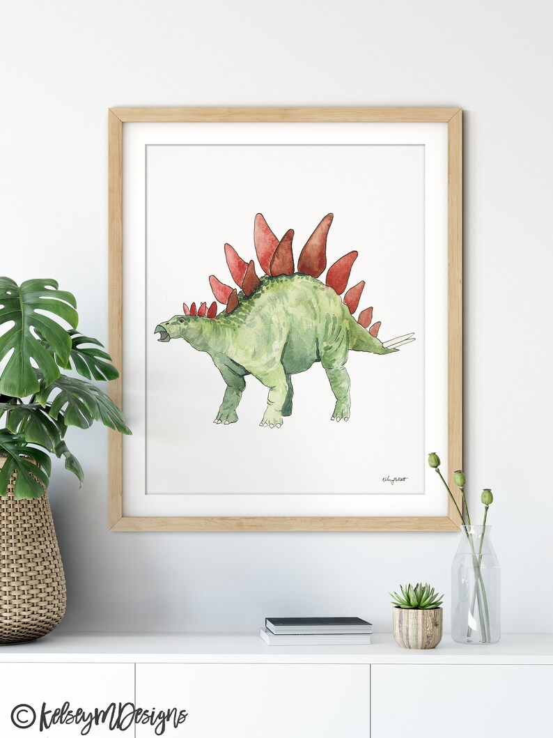 May include: Watercolor print of a green Stegosaurus with red plates, framed in a light wood frame. The artwork is on a white background. The artist's signature is visible. The print is displayed on a white surface.