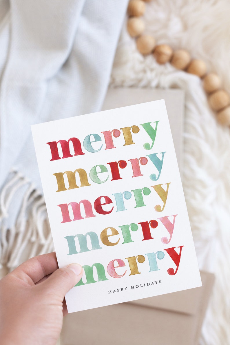 May include: A white card with the word "merry" repeated five times in different colors: red, gold, pink, blue, and gold. The word "merry" is in a bold, stylized font. The text "Happy Holidays" is printed in black at the bottom of the card.