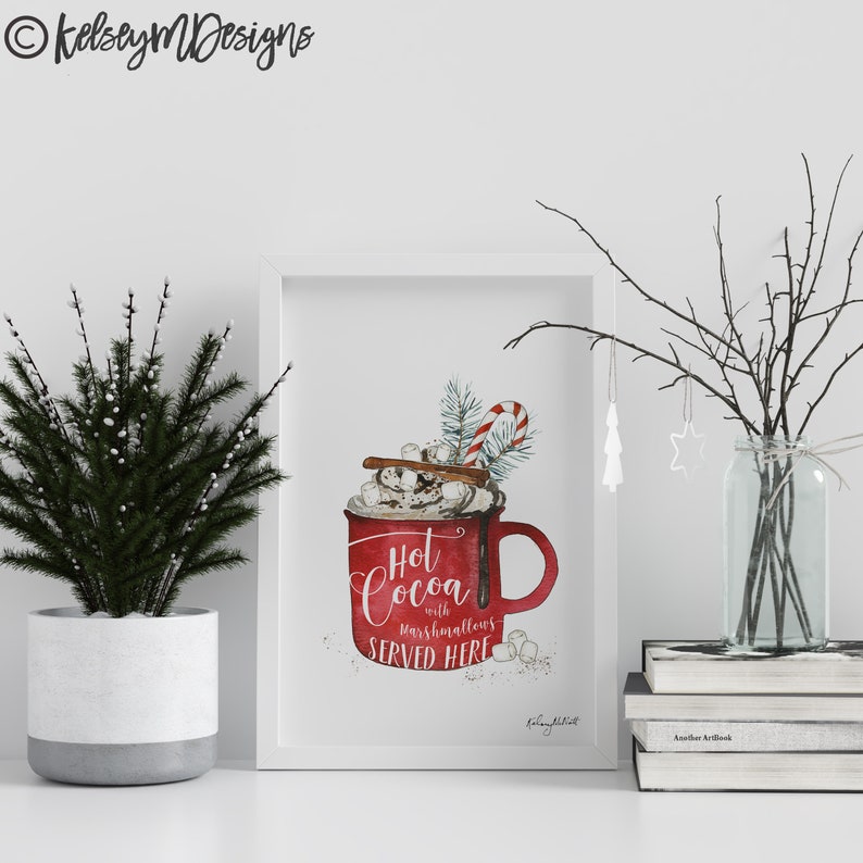 May include: White framed print with a watercolor illustration of a red mug filled with hot cocoa, marshmallows, whipped cream, cinnamon sticks, and a candy cane. The text reads "Hot Cocoa with marshmallows SERVED HERE".