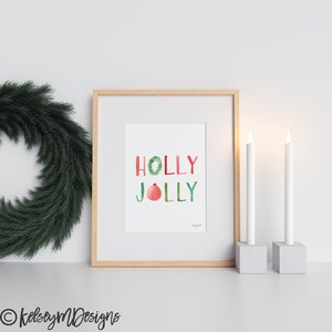 May include: A framed watercolor print with the words "HOLLY JOLLY" in colorful letters. A red and green holly wreath is incorporated into the word "HOLLY" and a red ornament is incorporated into the word "JOLLY". The print is displayed on a white wall with two white candles on a gray surface.
