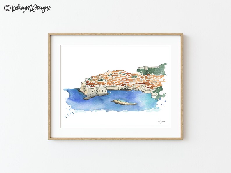 May include: Watercolor painting of a city on a peninsula with a blue ocean surrounding it. The city is made up of many buildings with red tile roofs. The painting is framed in a light wood frame.