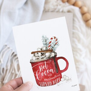 May include: A watercolor illustration of a red mug filled with hot cocoa, topped with whipped cream, marshmallows, and a cinnamon stick. The mug has the text "Hot Cocoa with marshmallows SERVED HERE" written on it.