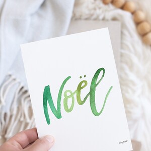May include: A white card with the word "No&euml;l" written in green watercolor script. The card is being held by a hand.
