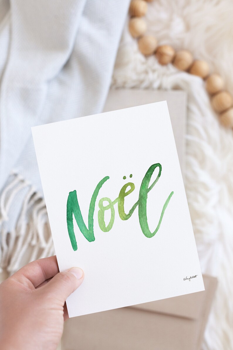 May include: A white card with the word "No&euml;l" written in green watercolor script. The card is being held by a hand.