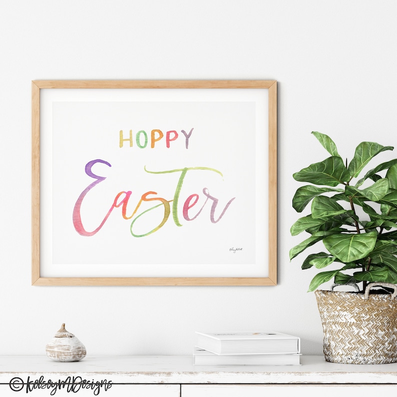 May include: A watercolor print with the text "Hoppy Easter" in rainbow colors. The print is framed in a light wood frame and is displayed on a white shelf with a small decorative object and a white book.