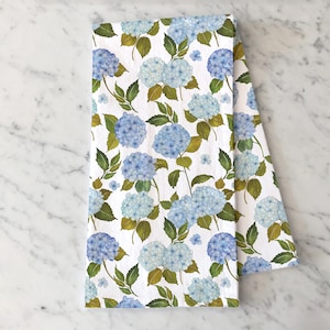Può includere: Un asciugamano da cucina bianco con un motivo floreale blu e verde. Il motivo presenta ortensie e foglie.