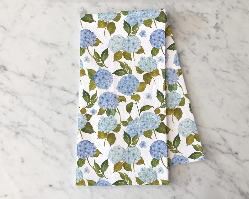 May include: A white kitchen towel with a blue and green floral pattern. The pattern features hydrangeas and leaves.