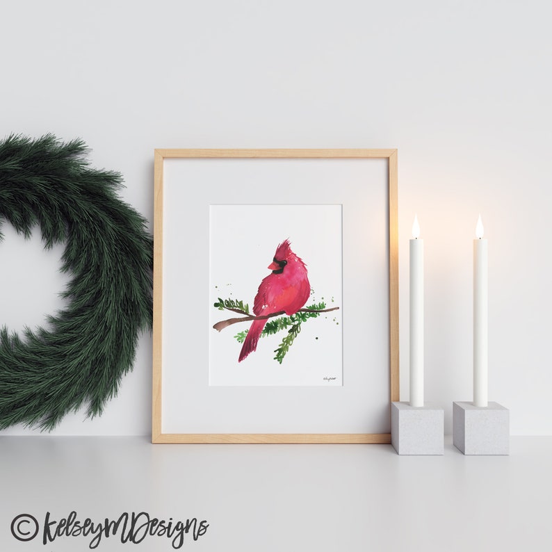 May include: A watercolor painting of a red cardinal perched on a branch with green leaves. The bird is facing to the left and has a black beak and black eyes. The background is white with a few splatters of paint.
