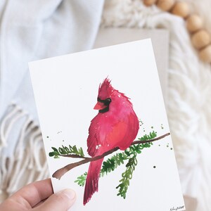 May include: A watercolor painting of a red cardinal perched on a branch with green leaves. The bird is facing to the left and has a black eye. The painting is on a white background.
