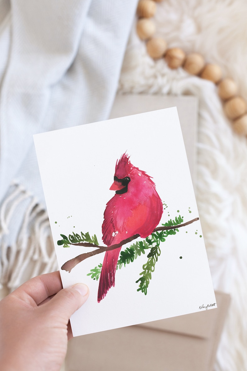 May include: A watercolor painting of a red cardinal perched on a branch with green leaves. The bird is facing to the left and has a black eye. The painting is on a white background.