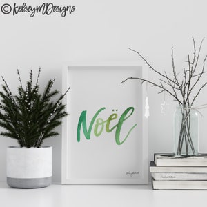 May include: A white framed print with the word "No&euml;l" written in green watercolor lettering. The print is sitting on a white surface with a small potted evergreen tree and a vase with branches.