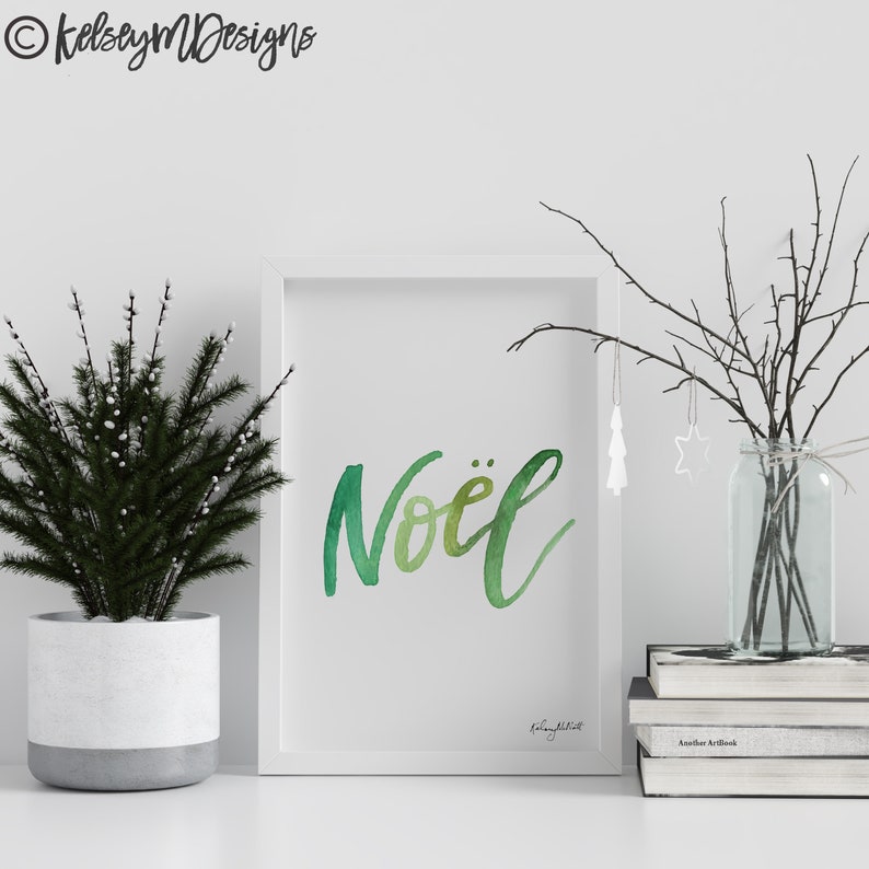 May include: A white framed print with the word "No&euml;l" written in green watercolor lettering. The print is sitting on a white surface with a small potted evergreen tree and a vase with branches.