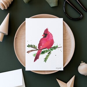May include: A watercolor painting of a red cardinal perched on a branch with green leaves. The bird is facing to the left and has a black beak and black eyes. The background is white.