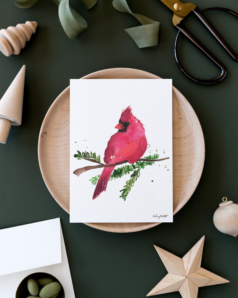 May include: A watercolor painting of a red cardinal perched on a branch with green leaves. The bird is facing to the left and has a black beak and black eyes. The background is white.