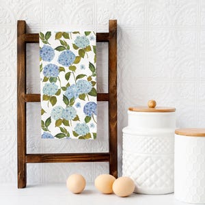 May include: A white kitchen towel with a blue and green floral pattern of hydrangeas. The towel is hanging on a wooden rack with two white ceramic canisters with wooden lids and three white eggs on a white surface.