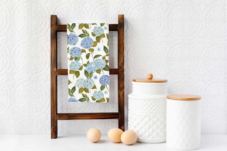 May include: A white kitchen towel with a blue and green floral pattern of hydrangeas. The towel is hanging on a wooden rack with two white ceramic canisters with wooden lids and three white eggs on a white surface.