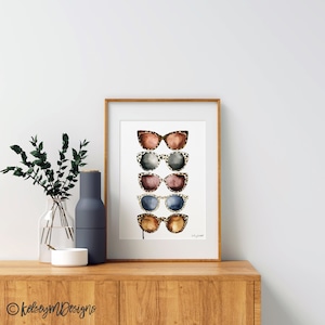 May include: A framed print of a watercolor painting of five pairs of sunglasses with leopard print frames. The sunglasses are stacked vertically and have different colored lenses. The print is on a white background and has the artist's signature in the bottom right corner.
