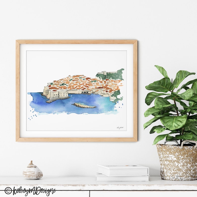 May include: A watercolor painting of a city on a peninsula with a blue sea surrounding it. The city is made up of many buildings with red tile roofs. The painting is framed in a light wood frame.