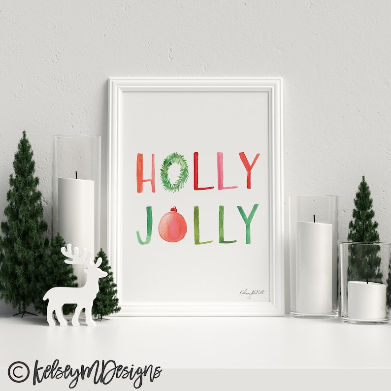 May include: A white framed print with the words "HOLLY JOLLY" in green, red, and pink watercolor lettering. A green wreath with a red ornament is incorporated into the lettering. The print is on a white background and has a white mat.
